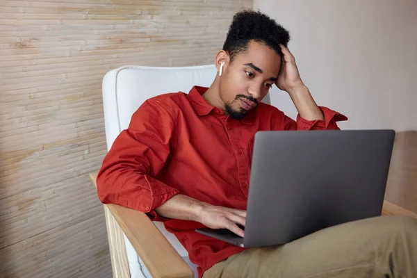 疲惫的年轻而卷曲的深色皮肤男性 留着胡子 手拿着头 凝视着笔记本电脑的屏幕 坐在家里的椅子上 — 图库照片