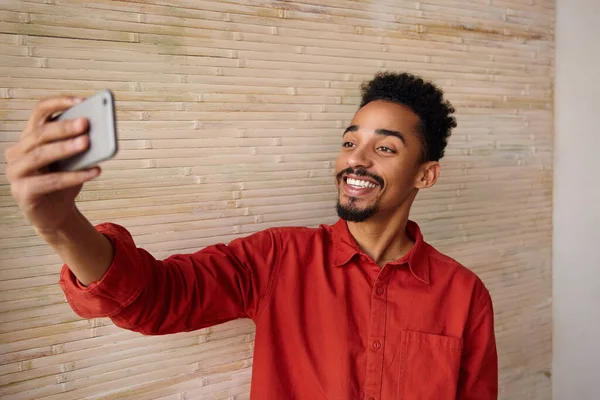 Pozitif, kısa saçlı, esmer, esmer, esmer, koyu tenli, akıllı telefonlu, elini kaldıran ve kameraya mutlu bir şekilde gülümseyen adam.