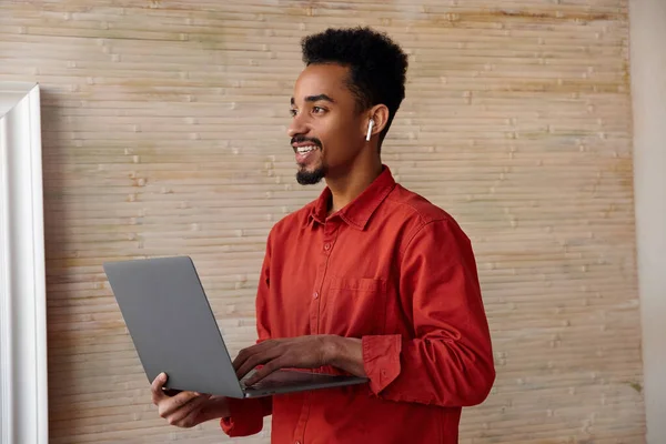 年轻迷人的短发黑发小伙子高举手提电脑 带着迷人的微笑向旁边看 站在米色的内部 侧面看 — 图库照片