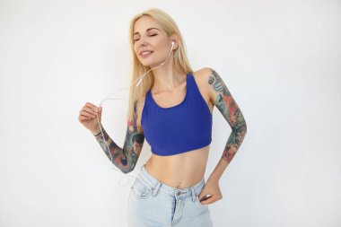 Müzik dinlerken ve dans ederken kulaklık takan, beyaz arka planda izole edilmiş, güzel, güzel, dövmeli, sarışın bir bayan.