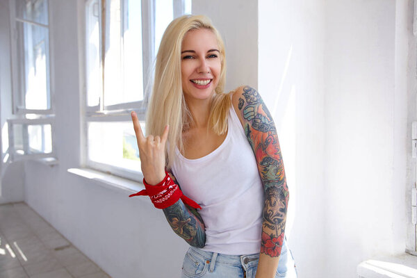 Крытое фото молодой блондинки с длинными волосами и татуировками, делающими рок-н-ролл вывеску с поднятой рукой и весело улыбающейся в камеру, позирующей перед большим окном
