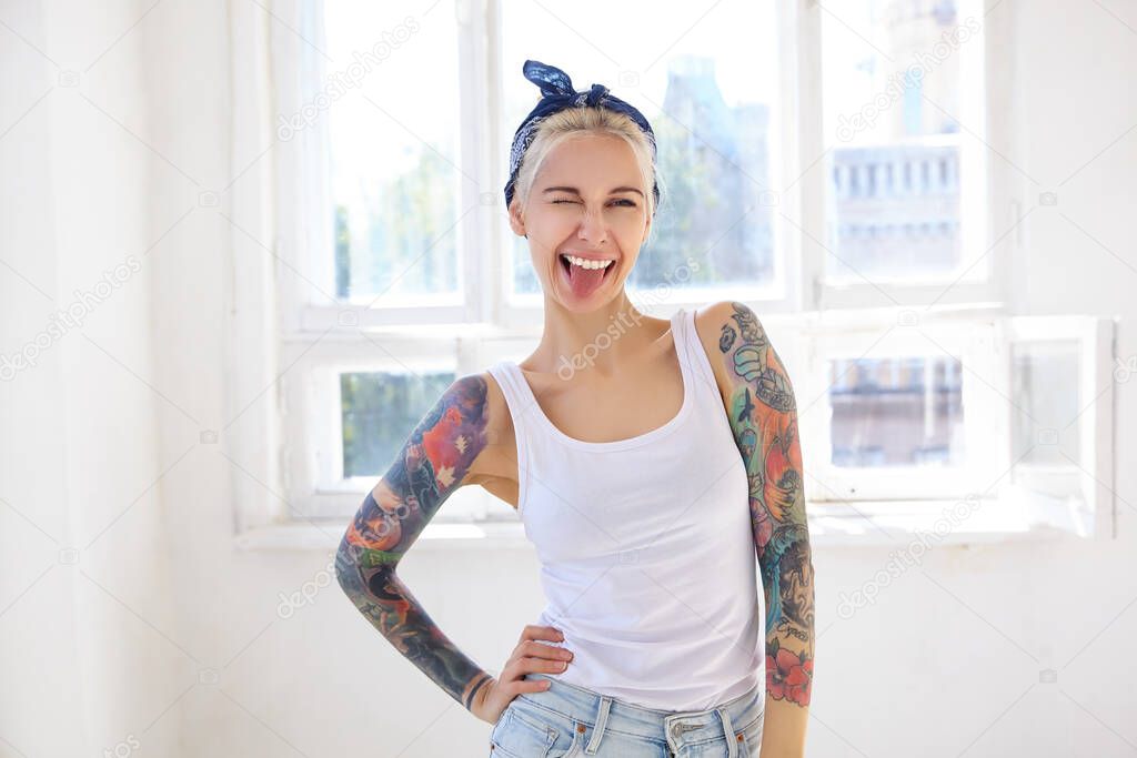 Joyful young attractive tattooed lady with casual hairstyle giving cheerfully wink at camera and showing tongue, keeping raised hand on waist while posing over bright studio