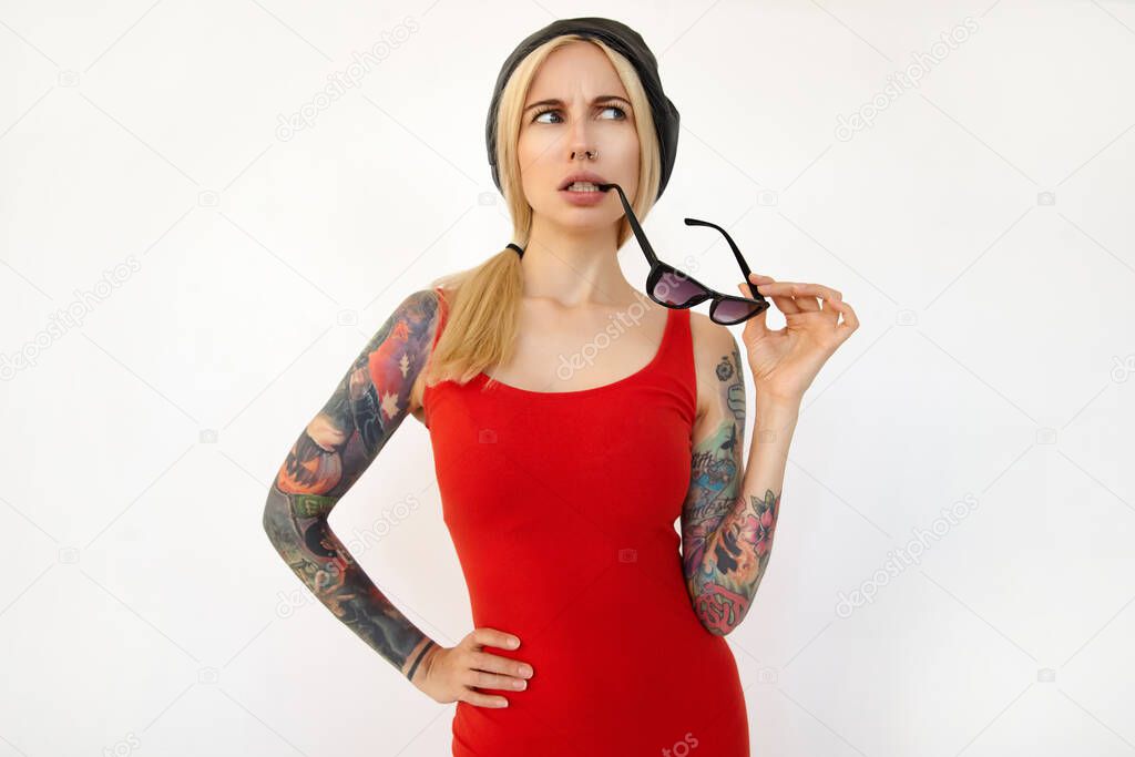 Bewildered young attractive tattooed long haired female with ponytail hairstyle biting worringly her sunglasses and frowning eyebrows while looking confusedly aside