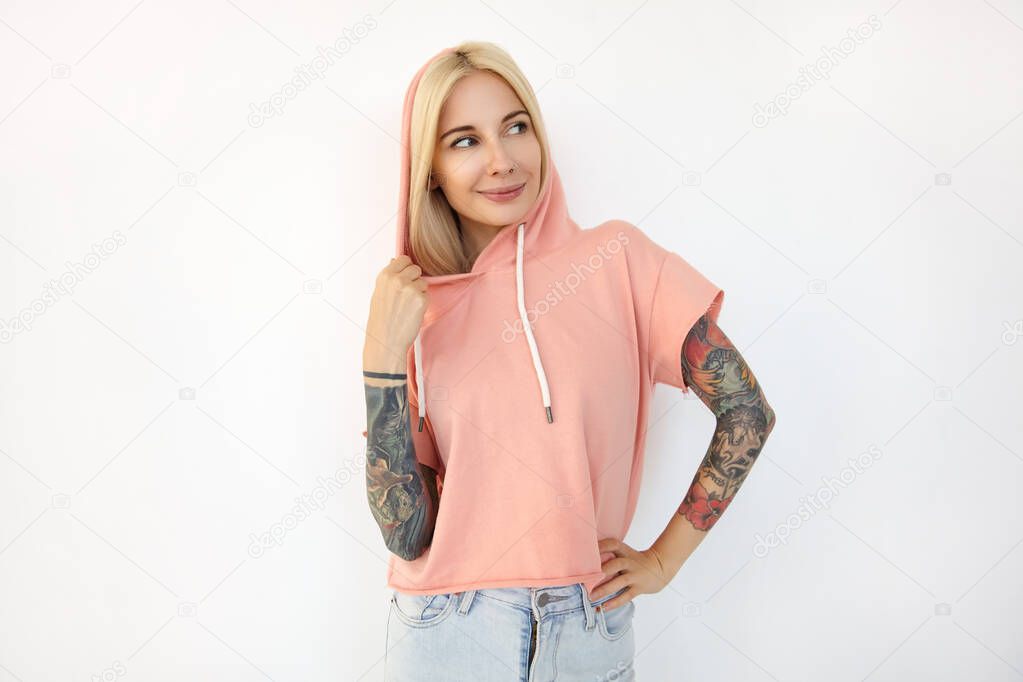 Positive young beautiful tattooed blonde woman dressed in sporty clothes keeping her hand on waist while looking gladly with pleasant smile, isolated over white background