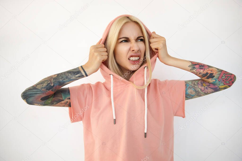 Displeased young attractive blonde lady with tattoos holding her hood with raised hands and grimacing her face while looking aside, isolated over white background