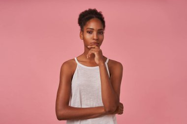 Çekici kahverengi saçlı, kıvırcık saçlı, koyu tenli bir kadının, kameraya dikkatlice bakarken gözlerini kısarak pembe arka planda poz verirken çekilmiş bir fotoğrafı.