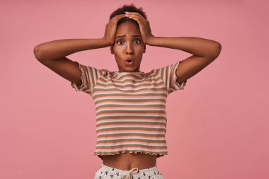 Shocked young pretty brunette dark skinned female clutching her head with raised hands while looking amazedly at camera, isolated over pink background clipart