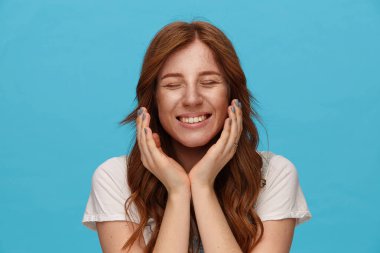 Güzel, güzel, kızıl saçlı, doğal makyajlı bir bayan ellerini yüzünün altında tutuyor ve mutlu bir şekilde gülümsüyor mavi arka planda soyutlanmış bir şekilde.