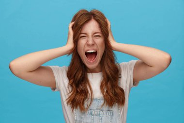 Stresli, kızıl saçlı, doğal makyajlı, ellerini kaldırmış, kaşlarını çatmış, ağzı açık bağıran, mavi arka planda poz veren genç bir kadın.