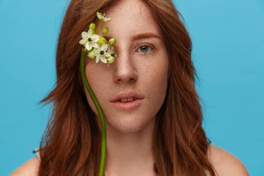 Güzel, yeşil gözlü kızıl saçlı, doğal makyajlı, sakin yüzüyle kameraya bakan, mavi arka planda poz verirken elinde çiçek dalı tutan bir kadın.