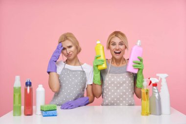 Mutsuz, beyaz kafalı temizlikçi kadın, heyecanlı sarışın kız arkadaşı pembe arka planda izole bir şekilde ellerini deterjanla kaldırırken üzgün bir şekilde kameraya bakıyor.