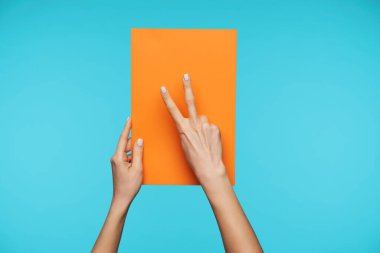 İki güzel kadının elleri A4 formatında bir kağıt tutarken ve zafer jestini parmaklarıyla gösterirken mavi arka planda izole edilmiş.