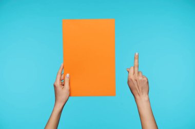 Young pretty fair-skinned woman's hands being raised while posing against blue background, keeping piece of paper in one hand and showing fuck gesture with another one
