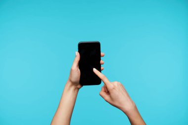 Güzel bir kadın beyaz manikürlü el kaldırdı akıllı telefonu tuttu ve işaret parmağıyla ekrandan geçti, mavi arka planda poz verdi.