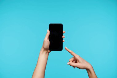 Modern cep telefonunu tutan ve mavi arka planda izole edilmiş, işaret parmağıyla siyah ekranda gösteren kaldırılmış bir kadının yatay görüntüsü.