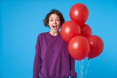 Tedirgin, genç, kıvırcık esmer kadın kameraya bakıyor geniş gözleri ve ağzı açık, mor kazak giyiyor ve mavi arka planda hava balonlarıyla poz veriyor.