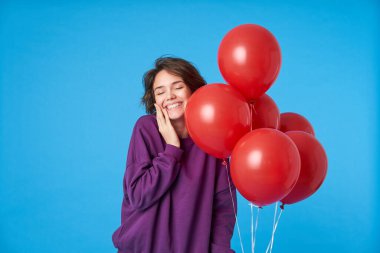 Güzel, güzel, koyu renk saçlı, doğal makyajlı bir kadın avuçlarını kaldırarak yanağına dokunuyor ve mavi arka planda mutlu bir şekilde gülümsüyor.