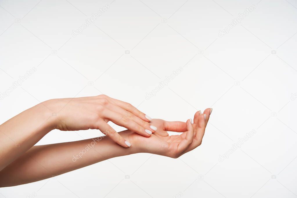 Beautiful young raised woman's hand with white manicure touching another one while moisturizing it with cream, isolated against white background