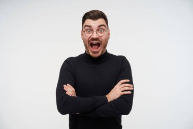 Heyecanlı, genç, sakallı esmer erkek göğsünde çapraz kollar ve kameraya hayret verici bir şekilde bakıyor geniş gözleri ve ağzı açık, beyaz arka planda duruyor.