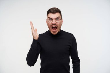 Kırgın, tıraşsız, kısa saçlı, kısa saçlı, ağzı açık, öfkeyle bağıran esmer bir adam beyaz arka planda dikilirken elini kaldırdı.