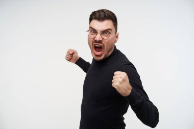 Kızgın, genç, yakışıklı, kısa saçlı, deli gibi bağıran ve yumruklarını sallayan esmer erkek. Beyaz arka planda izole edilmiş birine vuracak.