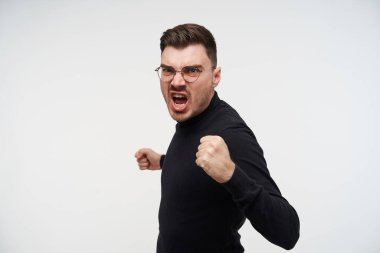 Stüdyodaki genç, huysuz, kısa saçlı, traşsız adamın dövüşe hazırlanırken yumruklarını kaldırdığı ve suratını asarak beyaz arka planda poz verdiği bir fotoğraf.