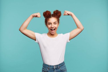 Aşırı neşeli, genç, kızıl saçlı kıvırcık kadın ellerini havada tutarak gücünü gösteriyor ve mavi arka planda izole bir şekilde neşeli bir şekilde kameraya bakıyor.
