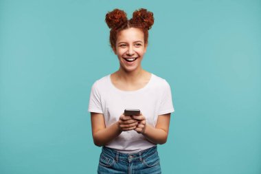 Kapalı alanda çekilen, kızıl saçlı, güzel bir kadın. Saçları topuz gibi, mutlu bir şekilde gülüyor. Akıllı telefonu elinde tutuyor, mavi arka planda soyutlanmış.