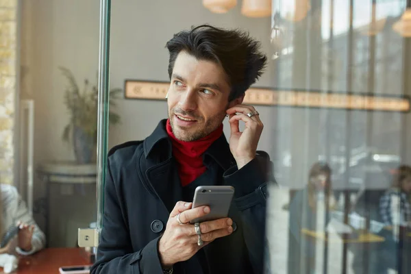 Close-up of young handsome dark haired unshaved male inserting earpiece while listening to music and looking positively aside, posing over cafe background