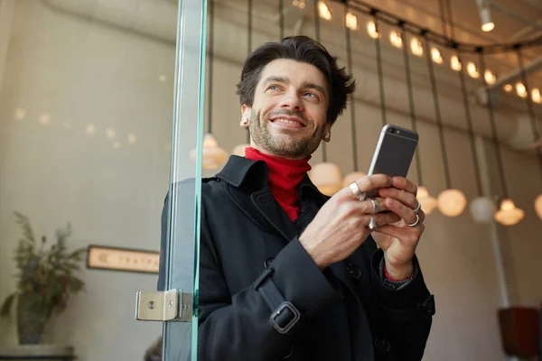 Agréable Jeune Beau Brun Aux Écouteurs Gardant Smartphone Regardant Joyeusement Photo De Stock