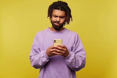 Kafası karışmış, koyu tenli, sakallı bir adam, sarı arka planda izole bir şekilde cep telefonunun ekranına bakarken yüzünü buruşturuyor.