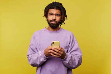 Şaşkın, kahverengi gözlü esmer adam akıllı telefonu kaldırmış ve sarı arka planda duran kameraya şaşkınlıkla bakıyor.