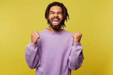 Aşırı neşeli, genç, koyu tenli esmer erkek mutlu bir şekilde bağırırken yüzünü buruşturuyor ve duygusal olarak ellerini kaldırıyor, sarı arka planda izole edilmiş.