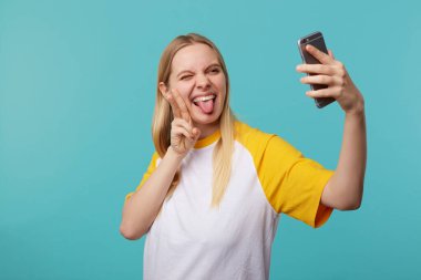 Kapalı alanda, sarışın, güzel bir kadının elini havada tutarken cep telefonunda selfie çekerken mavi arka planda poz verirken dalga geçtiği bir fotoğraf.