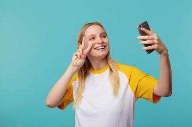 Çekici, beyaz saçlı, doğal makyajlı genç bir kadının portresi. Selfie çekerken gülümsüyor ve barış işareti ile el kaldırıyor. Mavi arka plan üzerinde izole edilmiş.