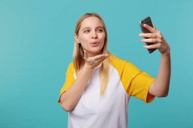 Pozitif, genç, sarışın, uzun saçlı, doğal makyajlı bir bayan. Mavi arka planda kendi fotoğrafını çekerken hava öpücüğü veriyor.
