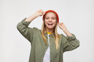 Neşeli, genç, güzel, beyaz saçlı, doğal makyajlı, mutlu bir şekilde kameraya bakan, geniş gülümsemeli, ellerini havada tutan, mavi arka planda soyutlanmış bir kadın.