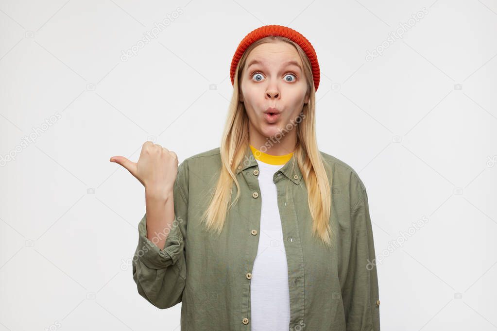 Dazed young attractive long haired blonde woman looking at camera with wide eyes opened and pointing aside with raised hand, standing over blue background