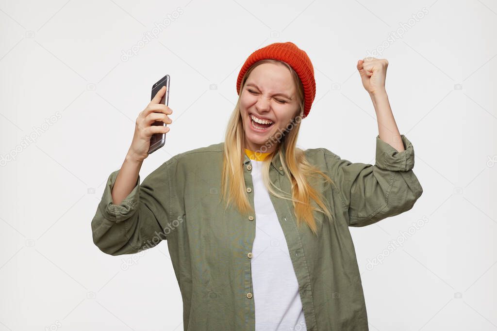 Studio photo of young attractive white-headed woman keeping her eyes closed while laughing happily with closed eyes, holding smartphone while posing over blue background