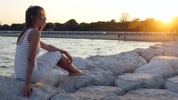 Молодая девушка в белом платье сидит на скалах на каменном пляже у моря на закате — стоковое видео