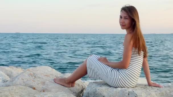 Jovem menina de vestido branco senta-se em rochas na praia de pedra no mar ao pôr do sol — Vídeo de Stock