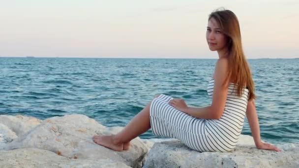 Молодая девушка в белом платье сидит на скалах на каменном пляже у моря на закате — стоковое видео