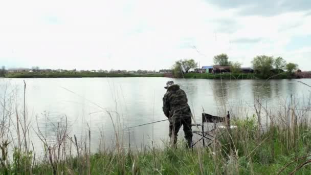 Largo tiro de Fishing Man en camuflaje sentado en silla con sombrero en un día nublado — Vídeos de Stock