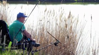 Yan görüş. Balıkçı, çalı çimenli göl kıyısında bir sandalyede oturur ve balık tutar.