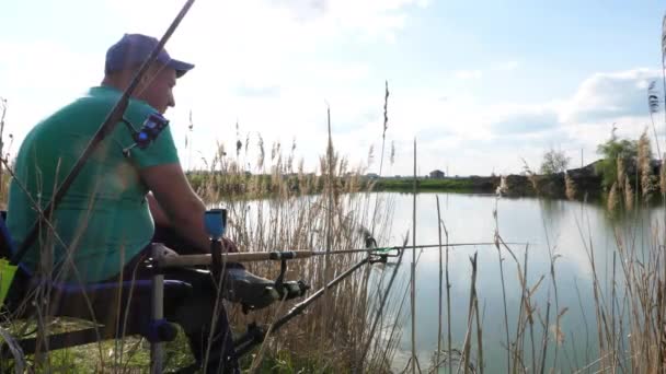 Pescador se sienta en el lago de hierba arbustiva orilla y la pesca. Día soleado. Vista lateral . — Vídeos de Stock