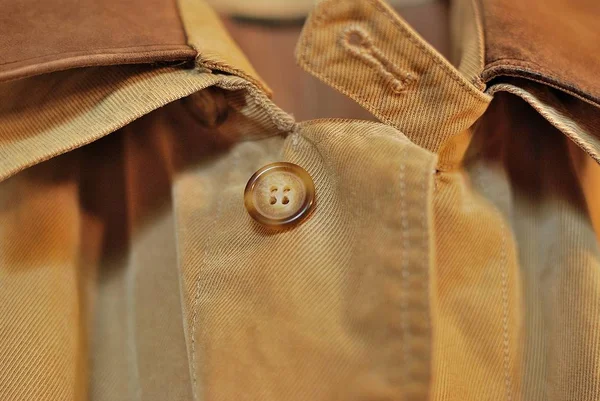 Şık bir yağmurluk üst düğmesi. Yağmurluğunun üst kısmı önümüzde. Düğme yakın çekimde. Kumaş dokusu, kum renkli tonları bütün zerafet altını çiziyor.
