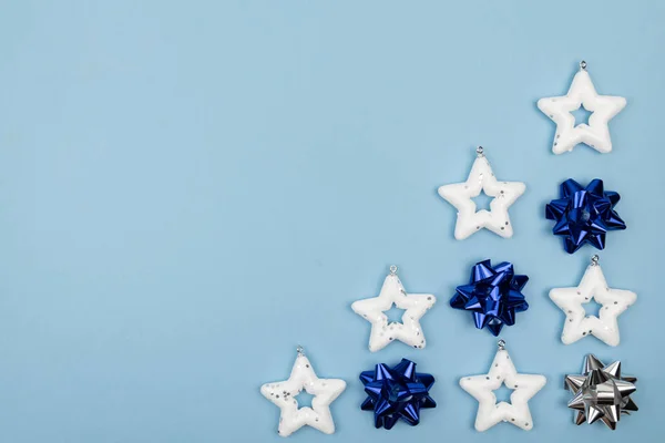 Tatil süslemeleri. Beyaz yıldızlardan oluşan yaratıcı kompozisyon - Noel ağacı oyuncakları, açık mavi arka planda klasik mavi ve gümüş fiyonklar. Üst manzara, yakın çekim. Boşluğu kopyala