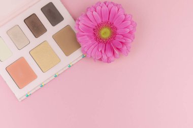 Pembe arka planda pembe gerbera çiçeğiyle çok renkli kozmetik makyaj malzemesi paleti. Pastel renk paleti, minimalizm, pastel pembe arka plan. Güzellik konsepti. Düz yatıyordu. Boşluğu Kopyala