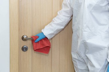 Koruyucu eldiven ve takım elbiseli kadın ya da erkek kırmızı mendil tutuyor ve metal kapı topuzunu temizliyor. Yeni tip solunum enfeksiyonuna karşı ofis dezenfeksiyonu Coronavirüs covid-19