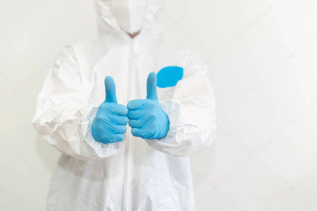 Doctor in white protective suit, medical mask and blue gloves shows a thumbs up. Epidemic, pandemic of coronavirus covid 19. Without face. Copy Space